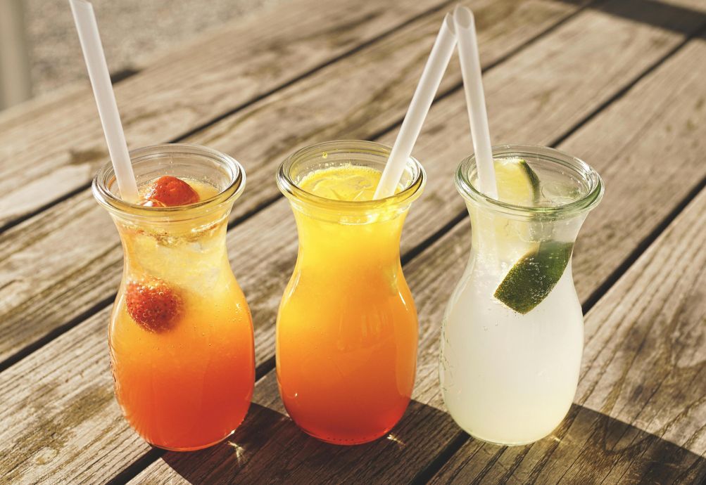 Close-up of three refreshing beverages with citrus, strawberries, and lime on a sunny wooden table.